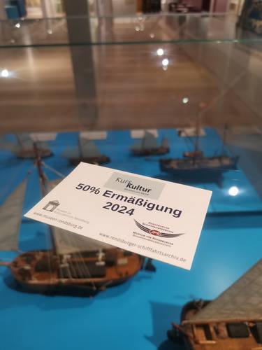 Ein Gutschein „50 % Ermäßigung 2024“ liegt auf einer Vitrine mit Schiffsmodellen.