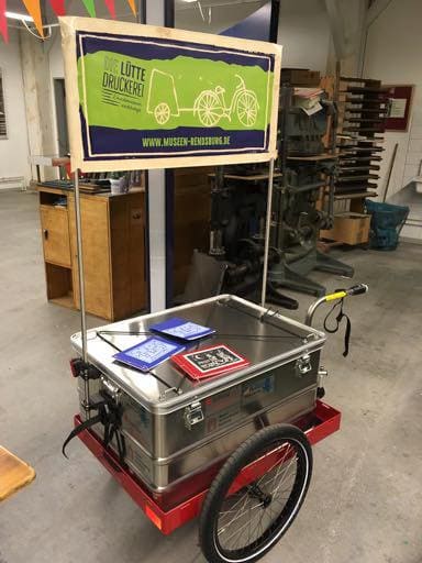 Fahrradanhänger der „lütten Druckerei“ mit einer Kiste voller Druckmaterial.